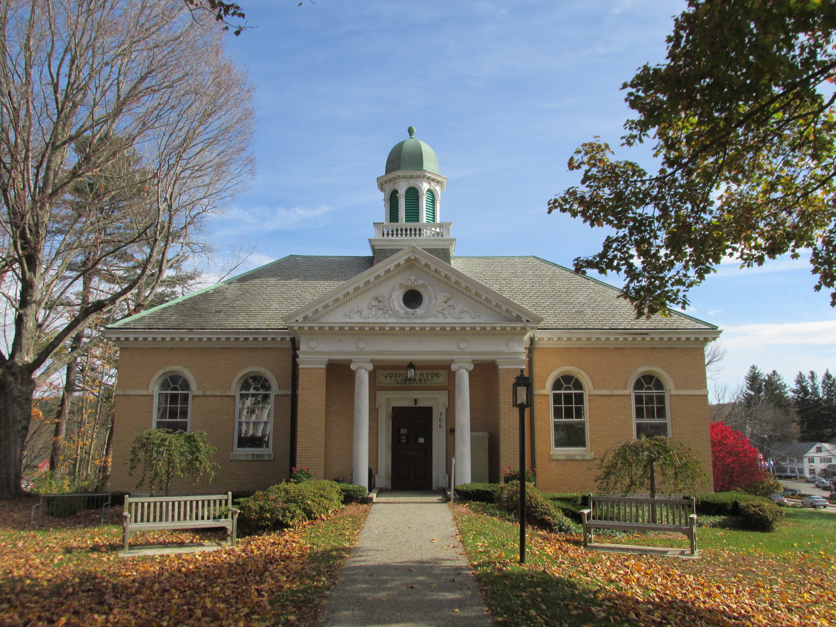 A small, tan brick library building with white columns and a green-domed cupola, surrounded by autumn trees and benches.