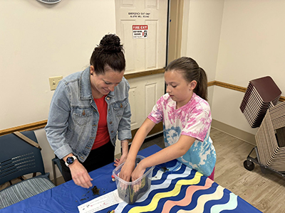 A woman and a child standing at the corner of a table with a bucket in front of them