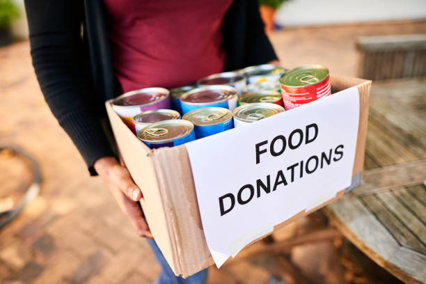 A person holding a cardboard box labeled "Food Donations" filled with canned goods. Background includes blurred outdoor wooden table, suggesting community support.