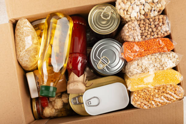 A cardboard box filled with assorted food items, including cans, oil, jars, and various packaged grains and legumes. The arrangement suggests organization and variety.