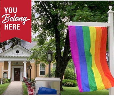 Pride flag hanging from a white post with a large house in the background