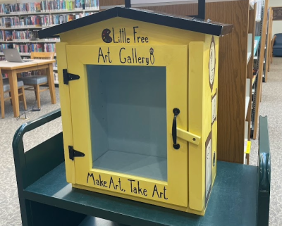 Yellow box labeled "Little Free Art Gallery" with glass front, in a library. Invitation to "Make Art, Take Art" creates a welcoming, creative vibe.