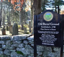 A historic cemetery with weathered gravestones and autumn trees. A sign reads "Old Burial Ground, Established c. 1740," conveying a respectful tone.