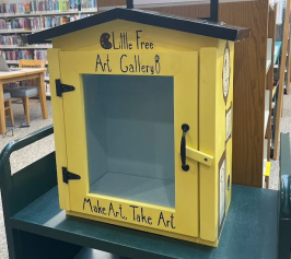 Yellow box labeled "Little Free Art Gallery" with glass front, in a library. Invitation to "Make Art, Take Art" creates a welcoming, creative vibe.