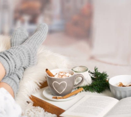 A person's legs in comfortable socks laying on a fuzzy pillow with a mug of hot chocolate and a book beside them.