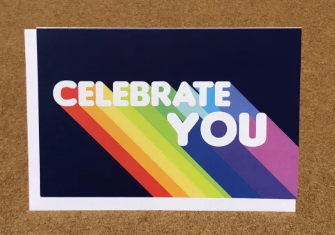 A card with the words "Celebrate You" in bold white letters over a rainbow-colored stripe background on a dark blue surface.
