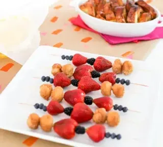 A white plate holds skewers with strawberries, blueberries, blackberries, and donut holes; a dish of sliced pastries and potted plants are visible in the background.