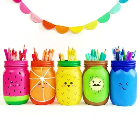 Five colorful jars painted as fruits—watermelon, orange, pineapple, kiwi, and blueberry—hold assorted pencils. A colorful garland hangs above, creating a playful, cheerful tone.