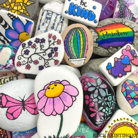 A collection of painted rocks featuring bright, colorful designs such as flowers, butterflies, a cactus, and positive words like "Be Kind." The overall tone is cheerful and uplifting.