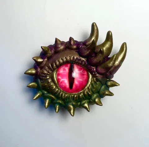 A vivid pink, reptilian eye with a vertical slit pupil, surrounded by a menacing array of golden spikes. The art conveys a sense of fantasy and danger.