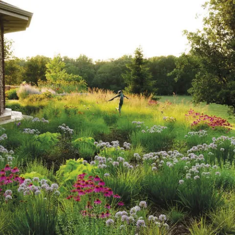 Lush garden with blooming purple and pink flowers, surrounded by tall grasses and trees. A small statue stands in the center, evoking a serene, peaceful mood.