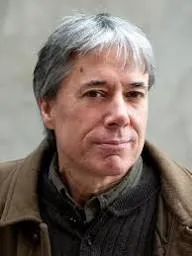 A man, Chuck Collins, with gray hair and a neutral expression looks at the camera. He's wearing a brown jacket and shirt. The background is a soft, blurred gray.