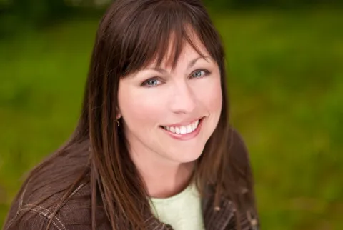 Smiling woman with long brown hair and bangs, wearing a brown jacket and green top, set against a blurred, lush green background.